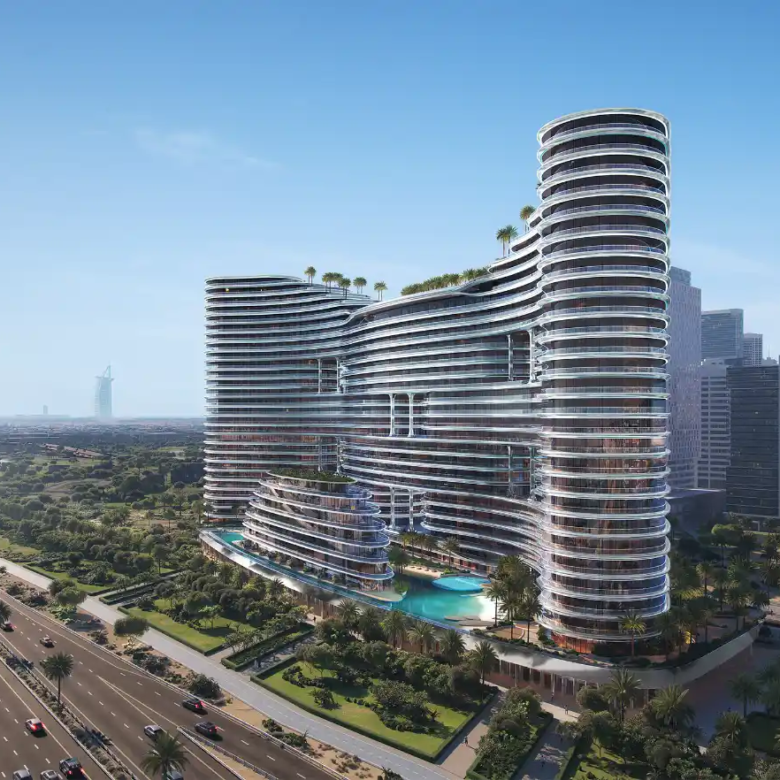 A modern, glass-clad high-rise with curved architecture—Binghatti Aquarise—features rooftop greenery and stands beside a busy multi-lane highway, surrounded by trees and cityscape under a clear blue sky.
