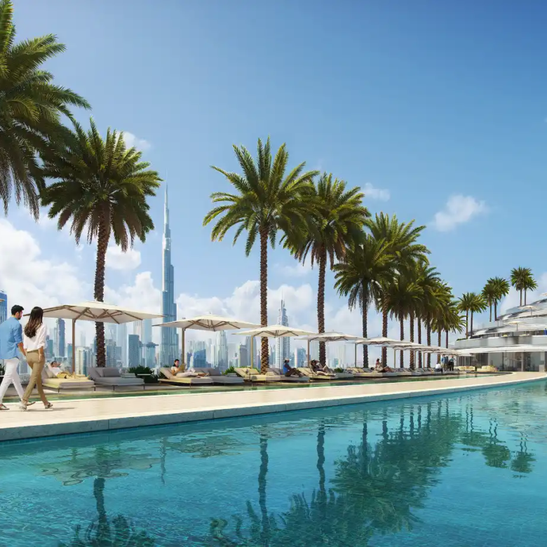 A couple walks beside the luxurious outdoor pool at Binghatti Aquarise, lined with palm trees and sun loungers, with a city skyline and the Burj Khalifa visible in the background under a bright blue sky.